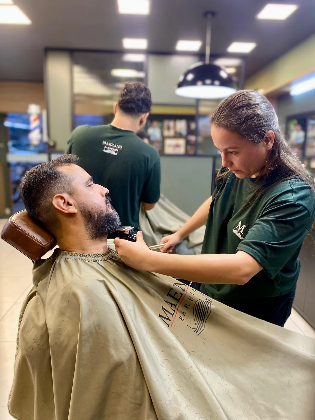 Barbeira fazendo a barba de um cliente na Maezano, com outro barbeiro atendendo e o salão ao fundo.